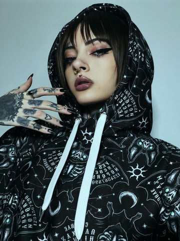A close-up of a model wearing a black hooded sweatshirt with a white celestial print of stars, moons, and occult symbols. The model has short dark hair, dramatic winged eyeliner, and a tattooed hand resting near their face.