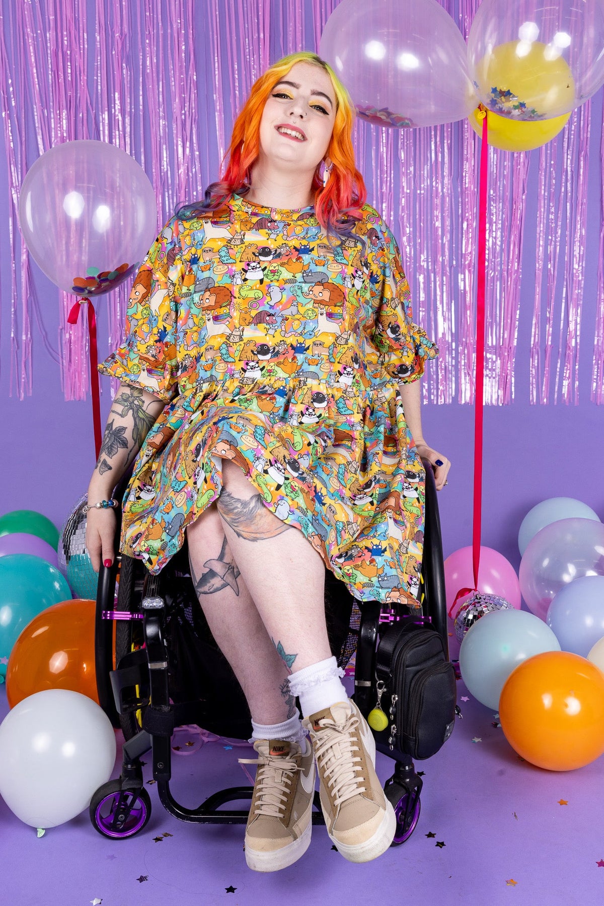 A person with orange and pink hair sits in a wheelchair wearing a colorful cartoon-patterned tunic shirt and beige sneakers, posed in a purple studio decorated with balloons and a metallic tinsel backdrop.
