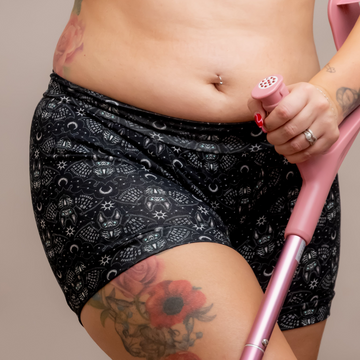 A close-up of a woman wearing a matching black and white celestial pattern activewear set, consisting of a sports bra and high-waisted shorts, holding a pink crutch against a solid beige background.
