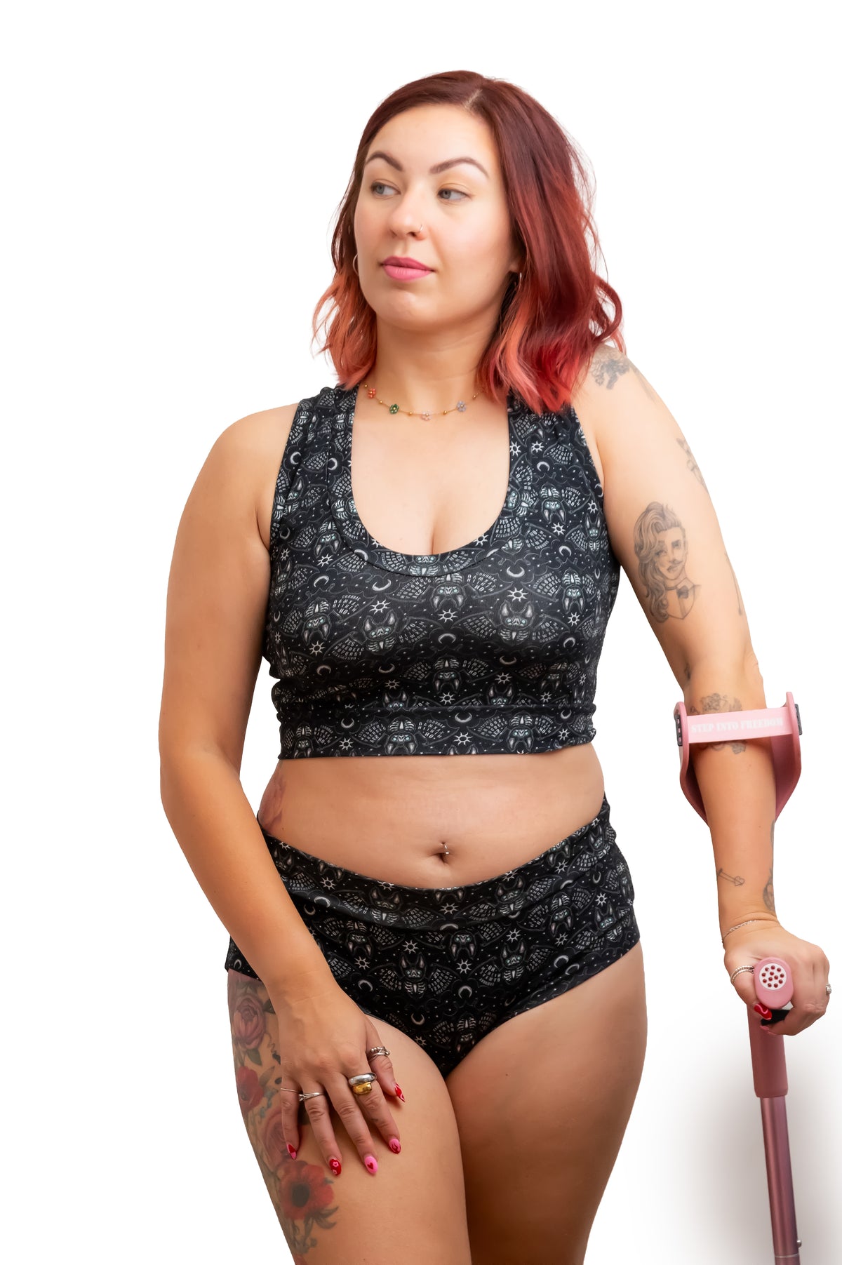A woman with red hair wearing a matching black and gray patterned sports bra and high-waisted brief set, holding a pink crutch against a white background.