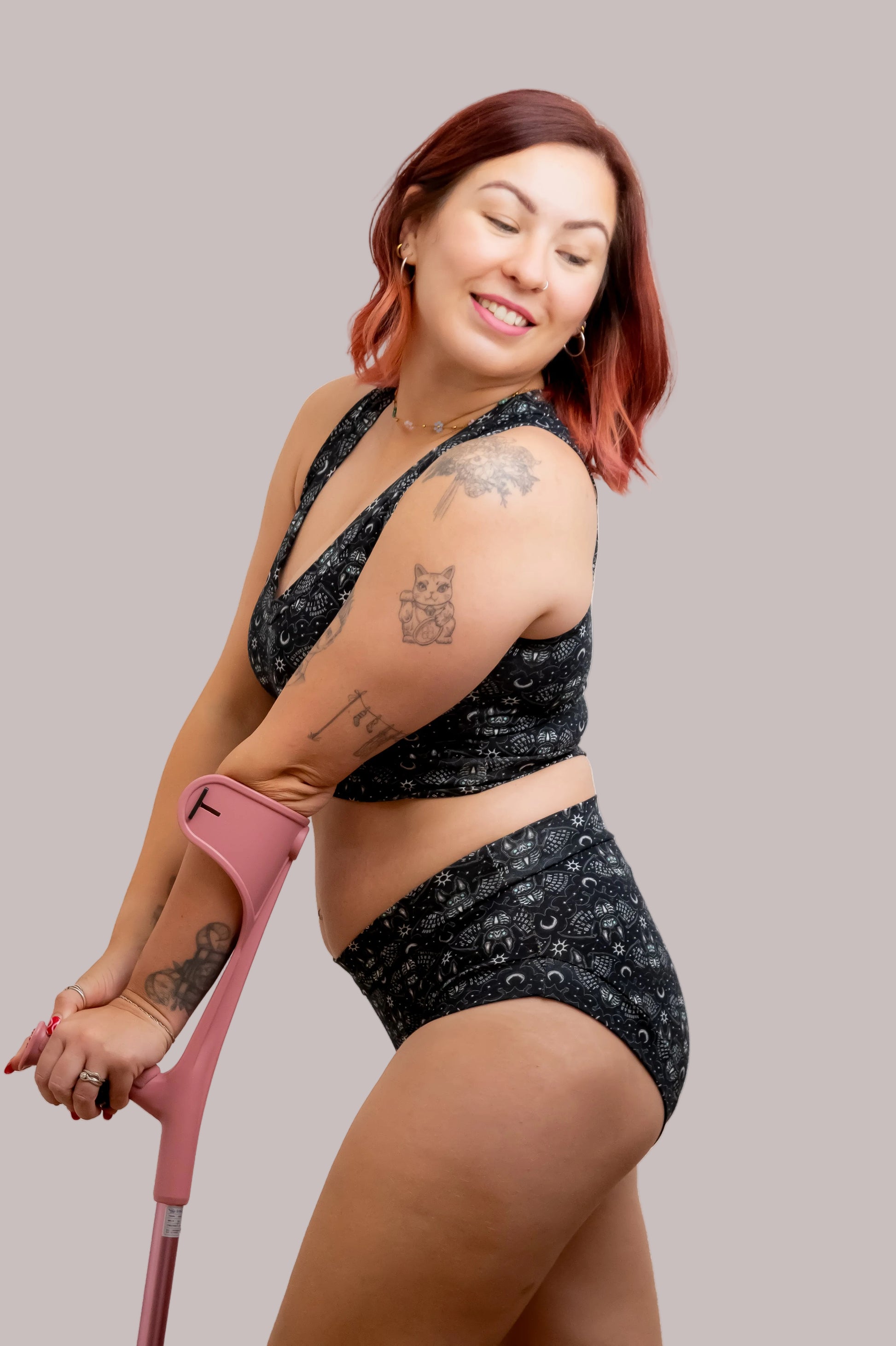 A woman with red hair posing with a pink underarm crutch, wearing a black and white patterned Ouija bra and matching high-waisted briefs against a solid white background.