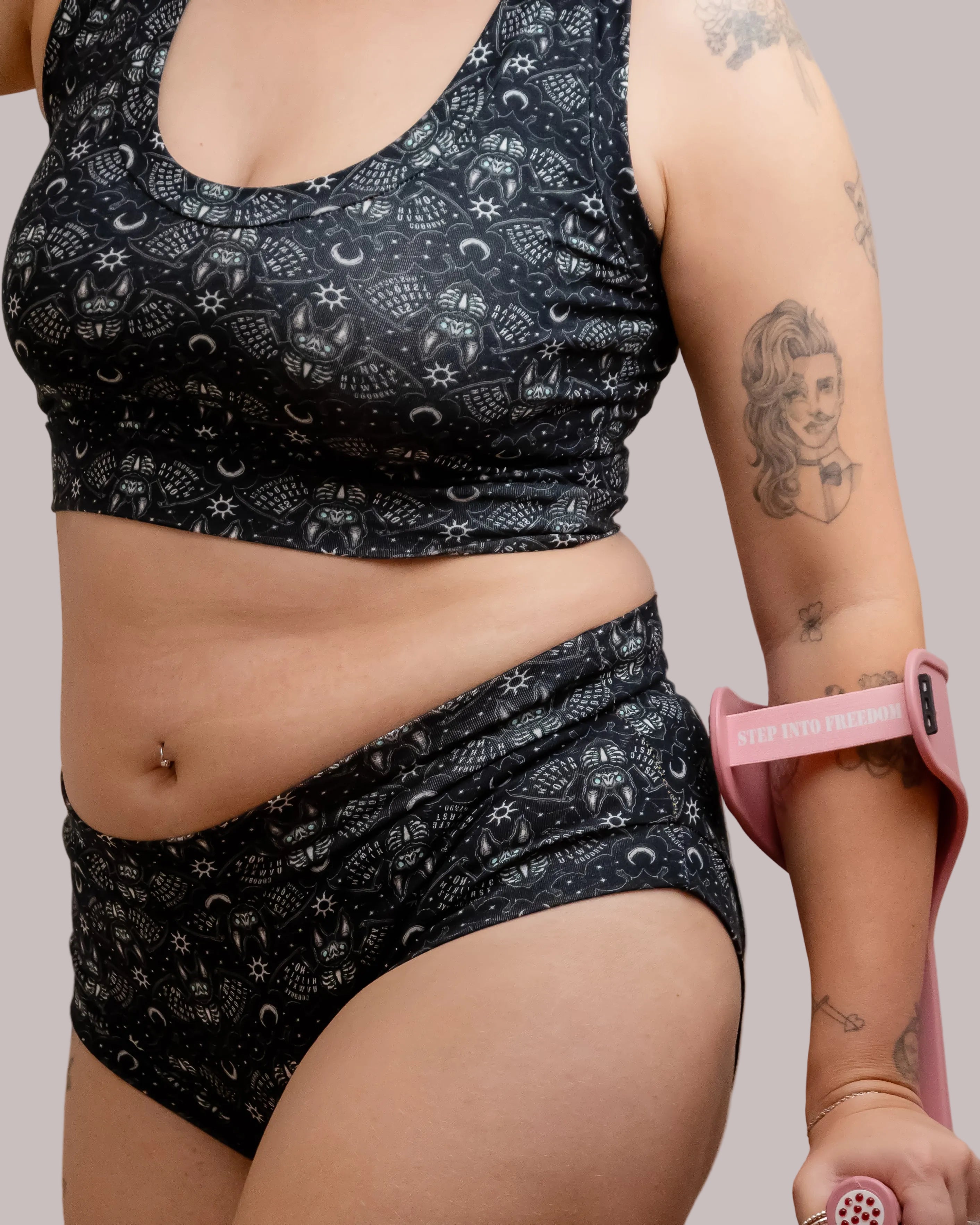A close-up of a person wearing a matching black and white Ouija board bats print underwear set, featuring a bralette and high-waisted briefs. The person is holding a pink crutch band around their upper arm.
