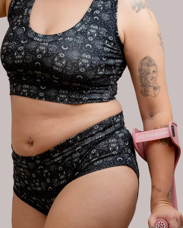 A close-up of a person wearing a matching black and white Ouija board bats print underwear set, featuring a bralette and high-waisted briefs. The person is holding a pink crutch band around their upper arm.