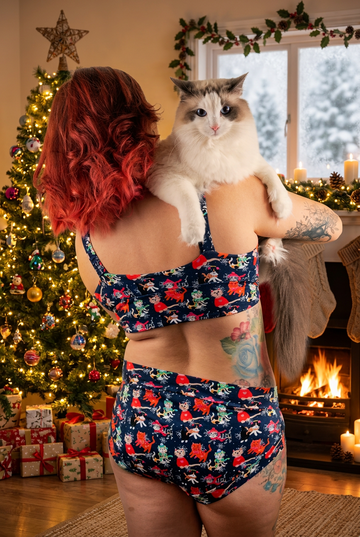 A woman with red hair is shown from behind wearing a navy blue two-piece swimsuit with a colorful floral print, while holding a fluffy white and gray cat on her shoulders against a solid white background.