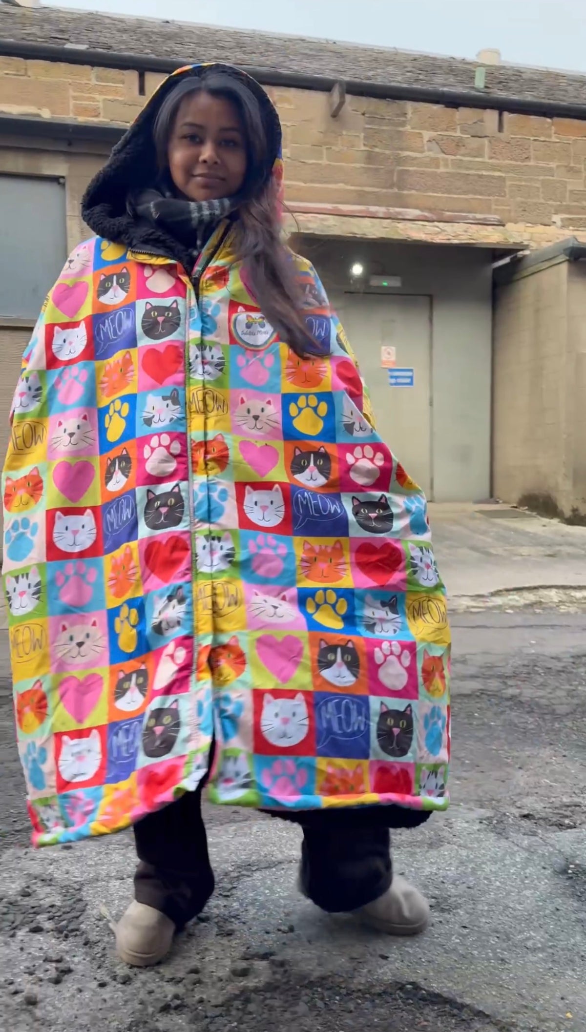 A person wearing a hooded rain poncho with a colorful patchwork print of cat faces, paw prints, and hearts, standing outdoors in front of a brick building.