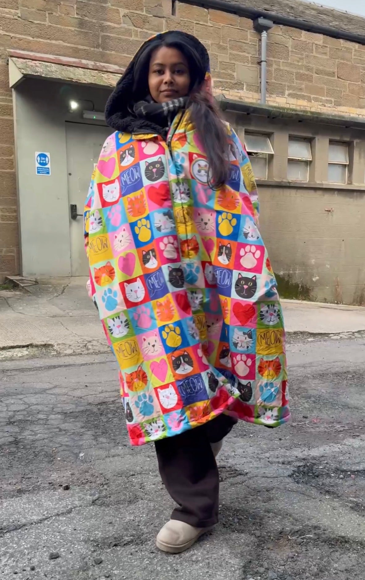 A person wearing a hooded poncho-style raincoat covered in a colorful patchwork cat-face pattern, standing outdoors on pavement in front of a brick building.