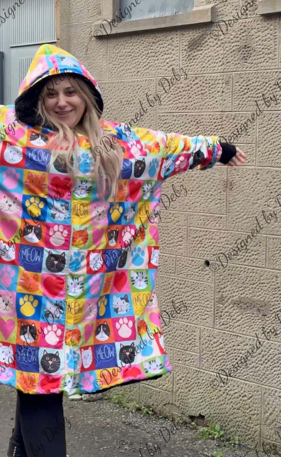 A woman wearing a hooded dry robe with a colourful patchwork design of cat and paw prints, standing outdoors against a textured concrete wall.