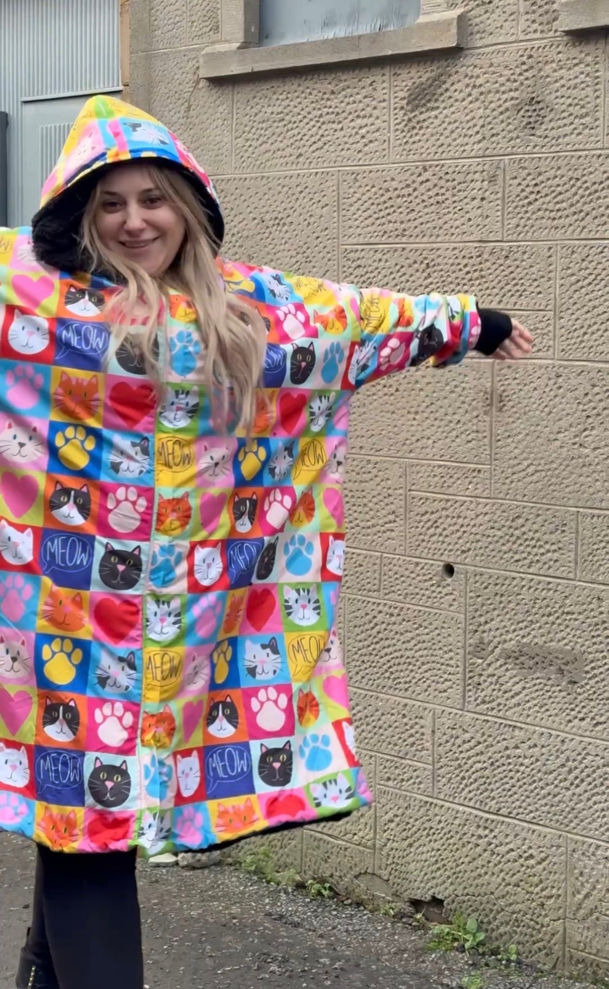A woman wearing a hooded dry robe with a colourful patchwork design of cat and paw prints, standing outdoors against a textured concrete wall.