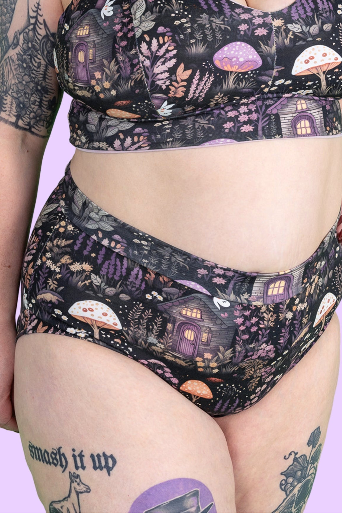A close-up of a person wearing a matching two-piece bikini with a whimsical forest print featuring purple mushrooms, fairy lights, and small houses on a black background, against a solid light purple backdrop.