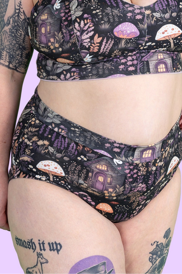 A close-up of a person wearing a matching two-piece bikini with a whimsical forest print featuring purple mushrooms, fairy lights, and small houses on a black background, against a solid light purple backdrop.