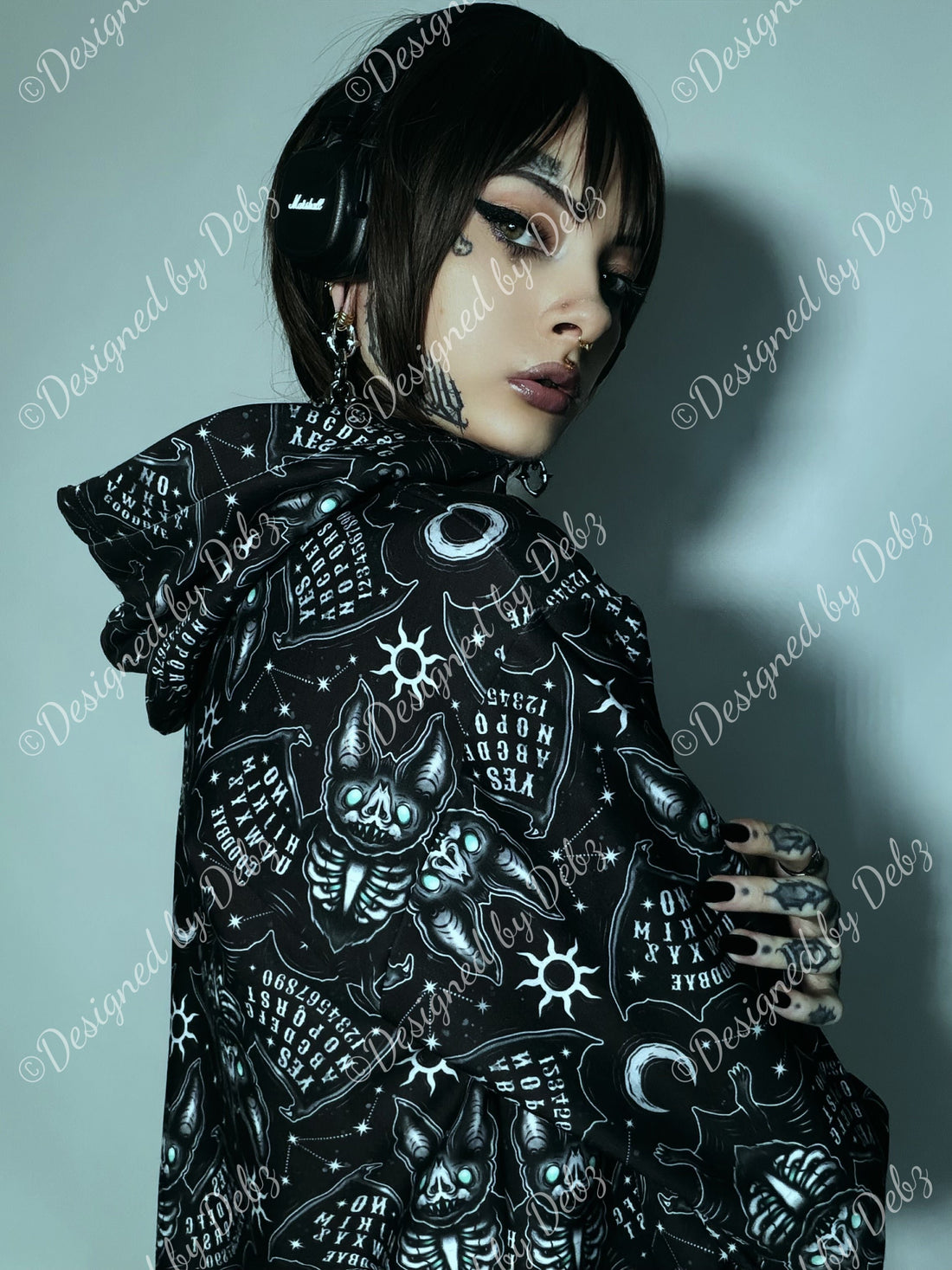 A model with short black hair, gothic makeup, and tattoos is shown from the back wearing a black hooded sweatshirt covered in a detailed white gothic illustration print. She is wearing black over-ear headphones against a cool gray studio background.