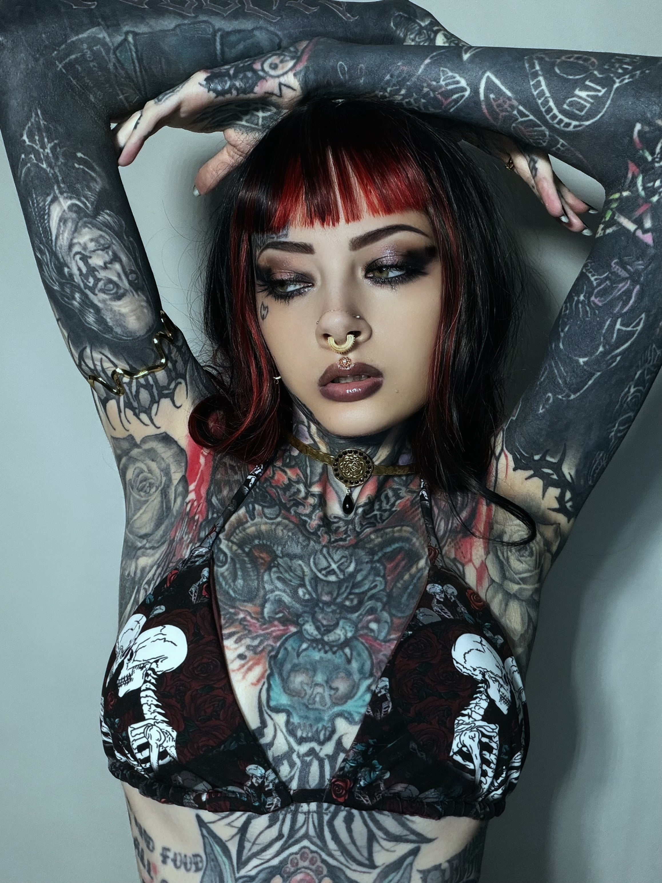 A tattooed woman with red-and-black hair poses with her arms raised, wearing a sheer black mesh bra top with white skeleton prints and a dark red underlayer.