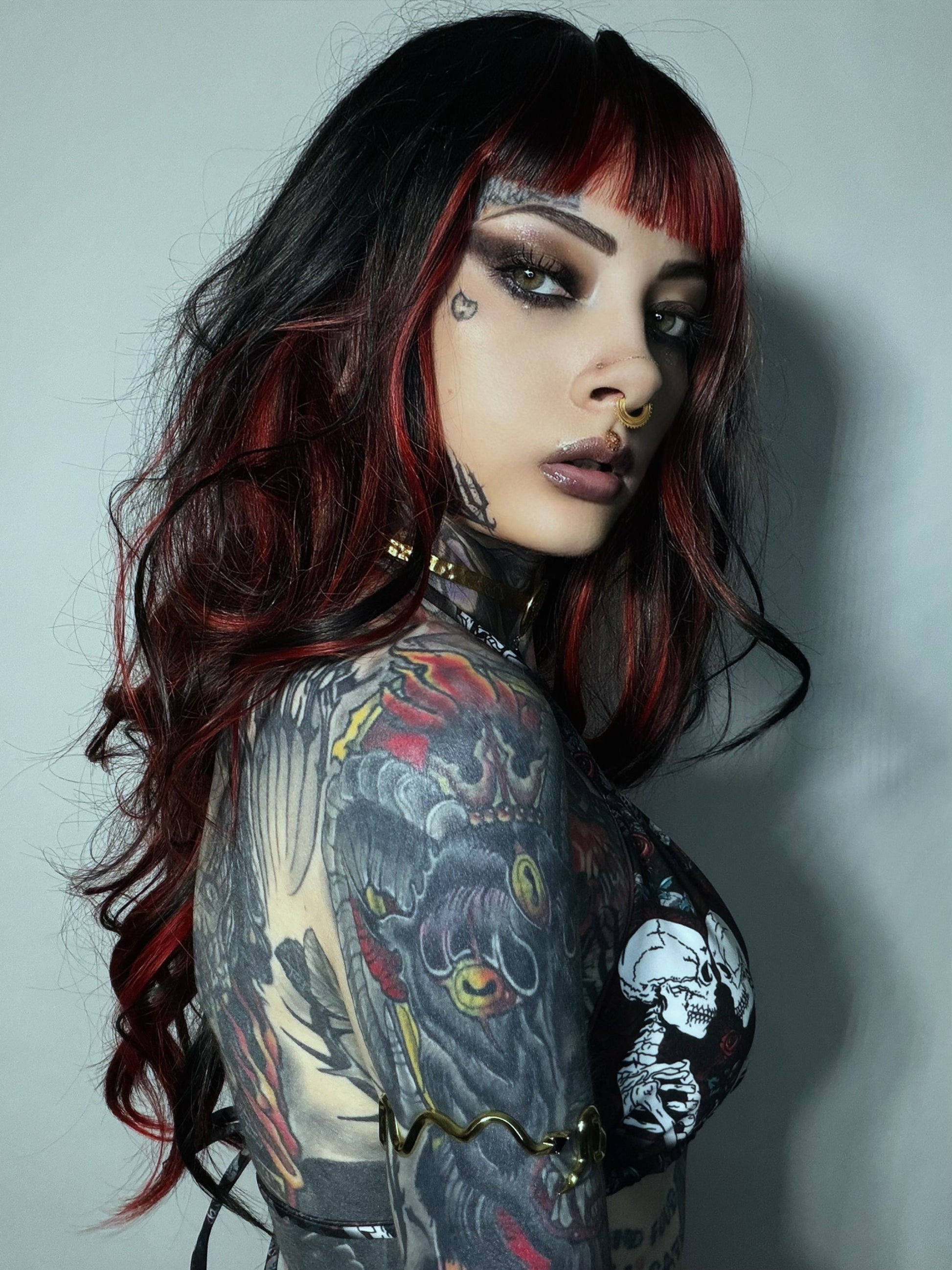 A woman with long black hair featuring vibrant red highlights and bangs, wearing a black graphic top with skull designs. She has extensive tattoos on her arms and neck, and is posed against a plain gray wall.
