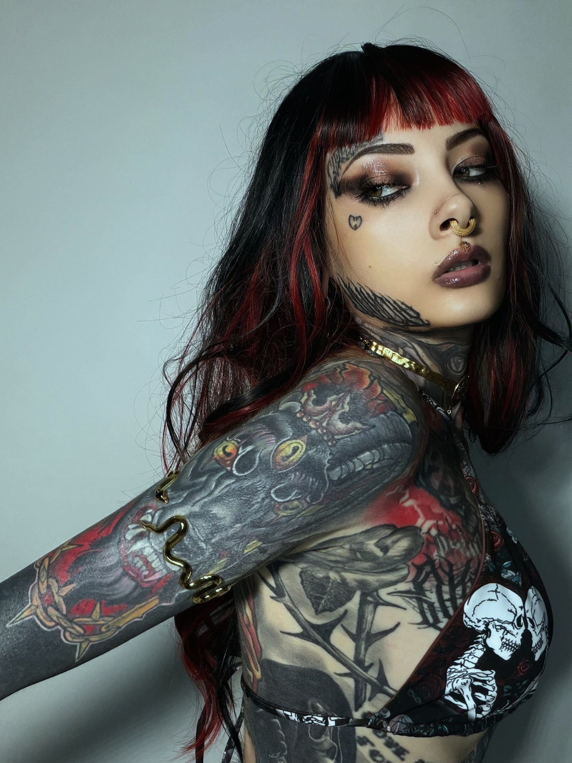 A close-up portrait of a woman with extensive black and gray tattoos on her arms and neck, featuring red and black hair, wearing a black bikini top with a white skeleton print against a plain gray background.