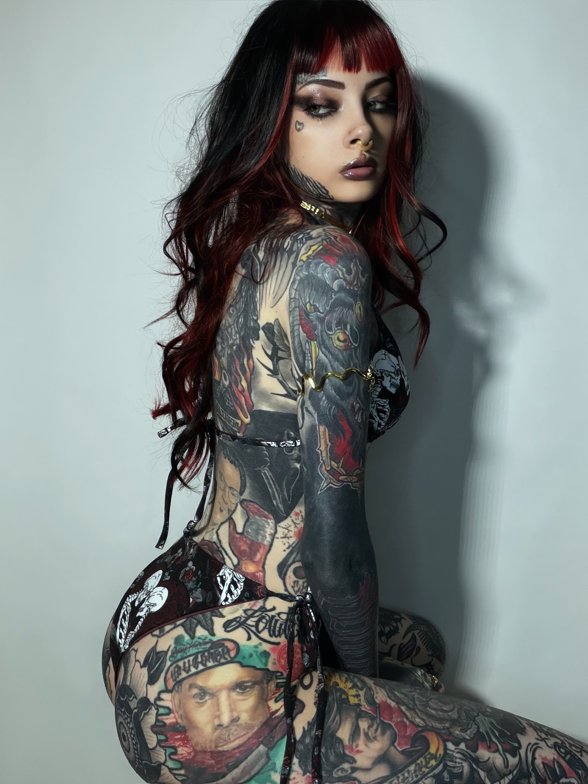 A tattooed woman with long black and red hair poses in a black lace-up bodysuit against a plain light gray wall.