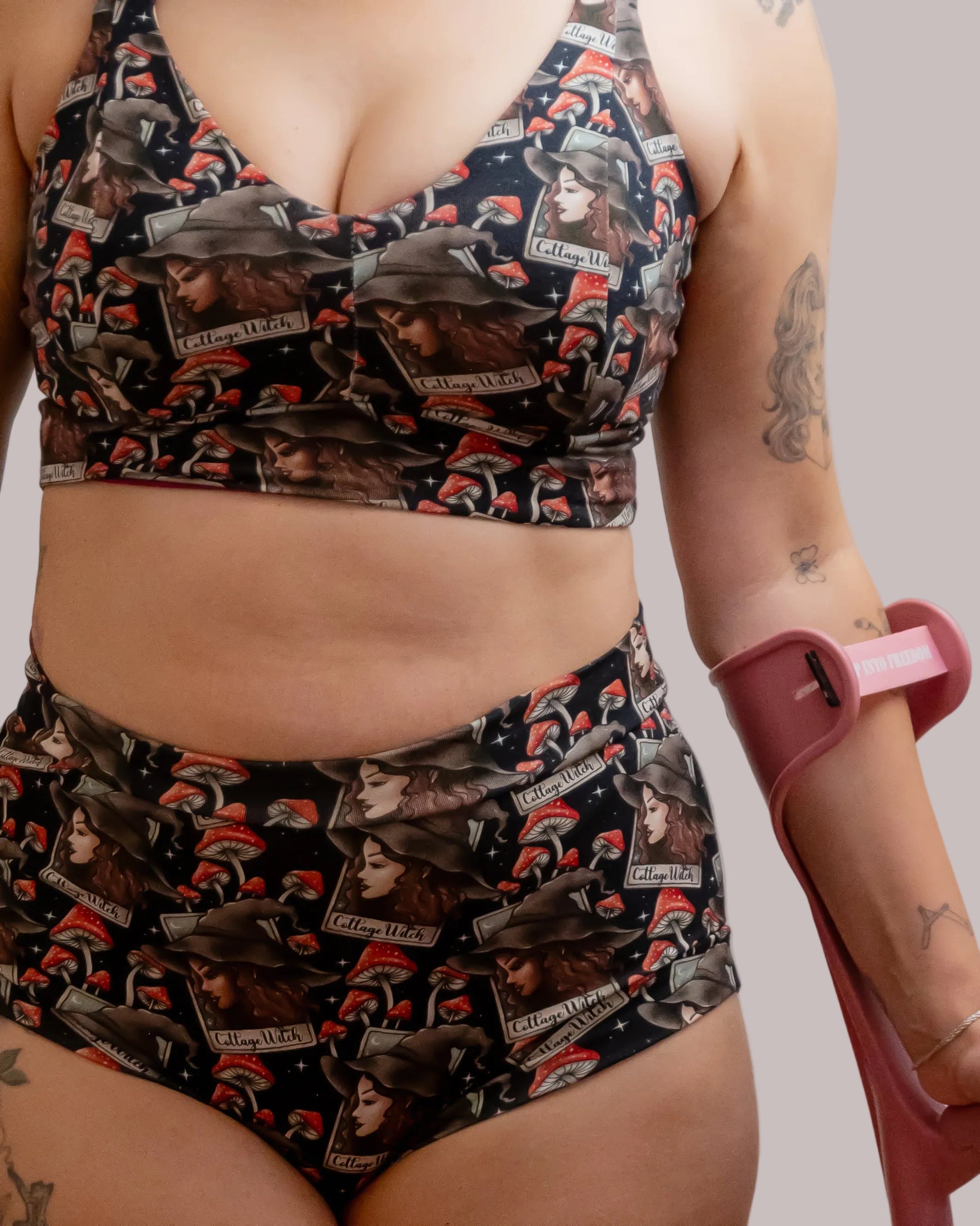 A close-up of a model wearing a matching high-waisted underwear set with a cottage witch, featuring black, red, and sepia-toned illustrations of black and white witches, toadstools and stars. The model is holding a pink crutch against a plain light gray background.