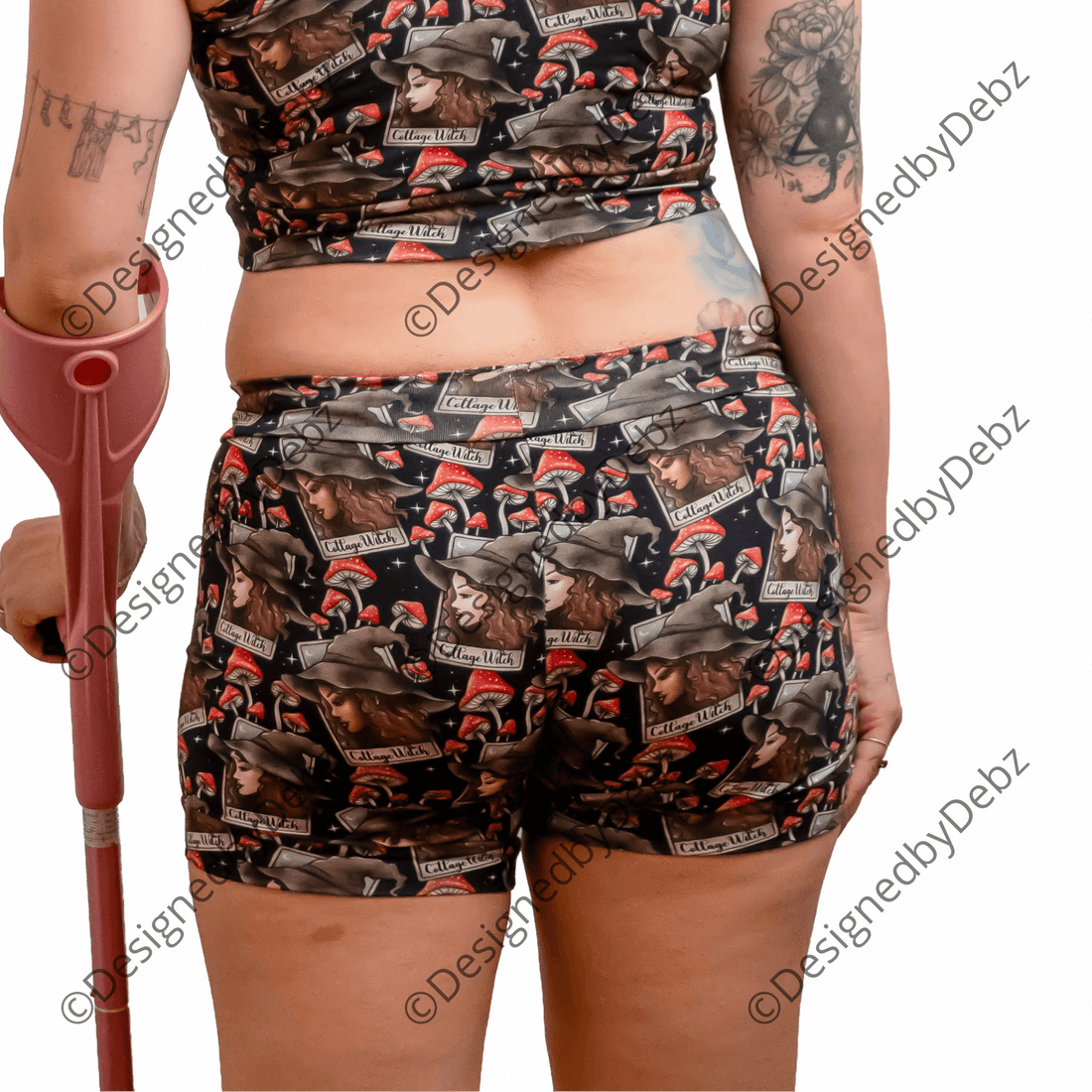 A woman with red hair is shown from behind wearing a matching camouflage print sports bra and shorts set, holding a pink crutch against a solid white background.