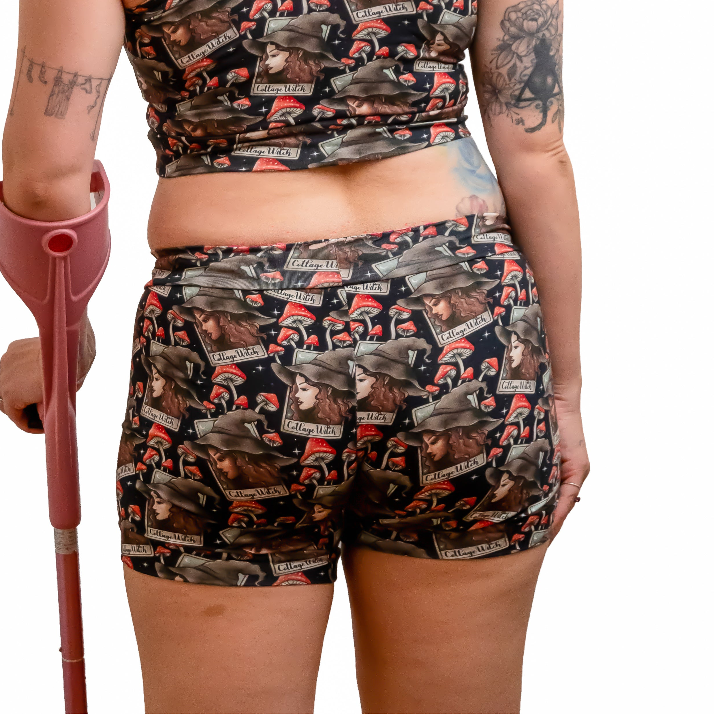 A woman with red hair is shown from behind wearing a matching camouflage print sports bra and shorts set, holding a pink crutch against a solid white background.