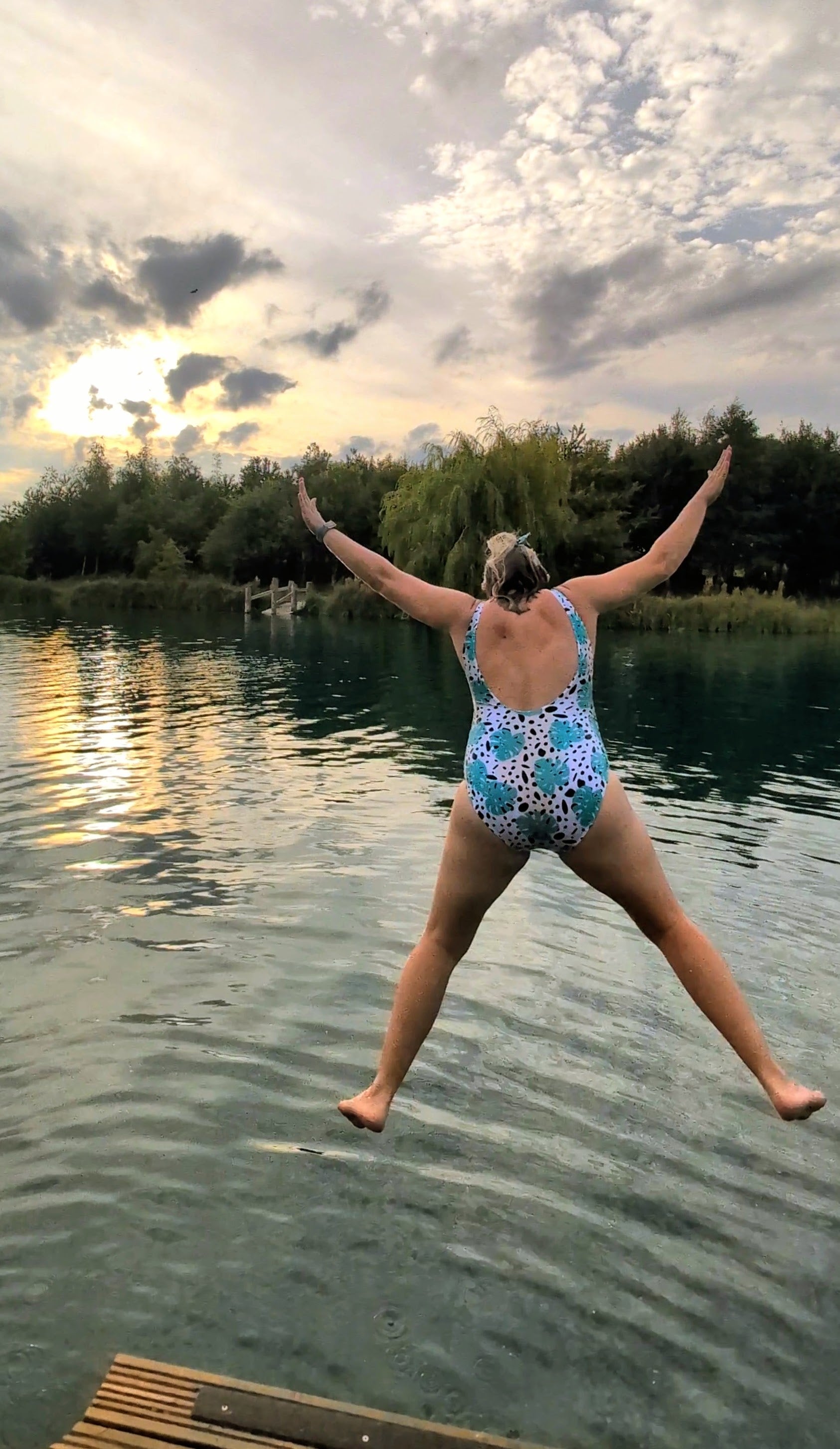 A woman wearing a blue and white patterned one-piece swimsuit jumps from a wooden dock into a calm lake, viewed from behind as she spreads her arms wide against a cloudy sunset sky.