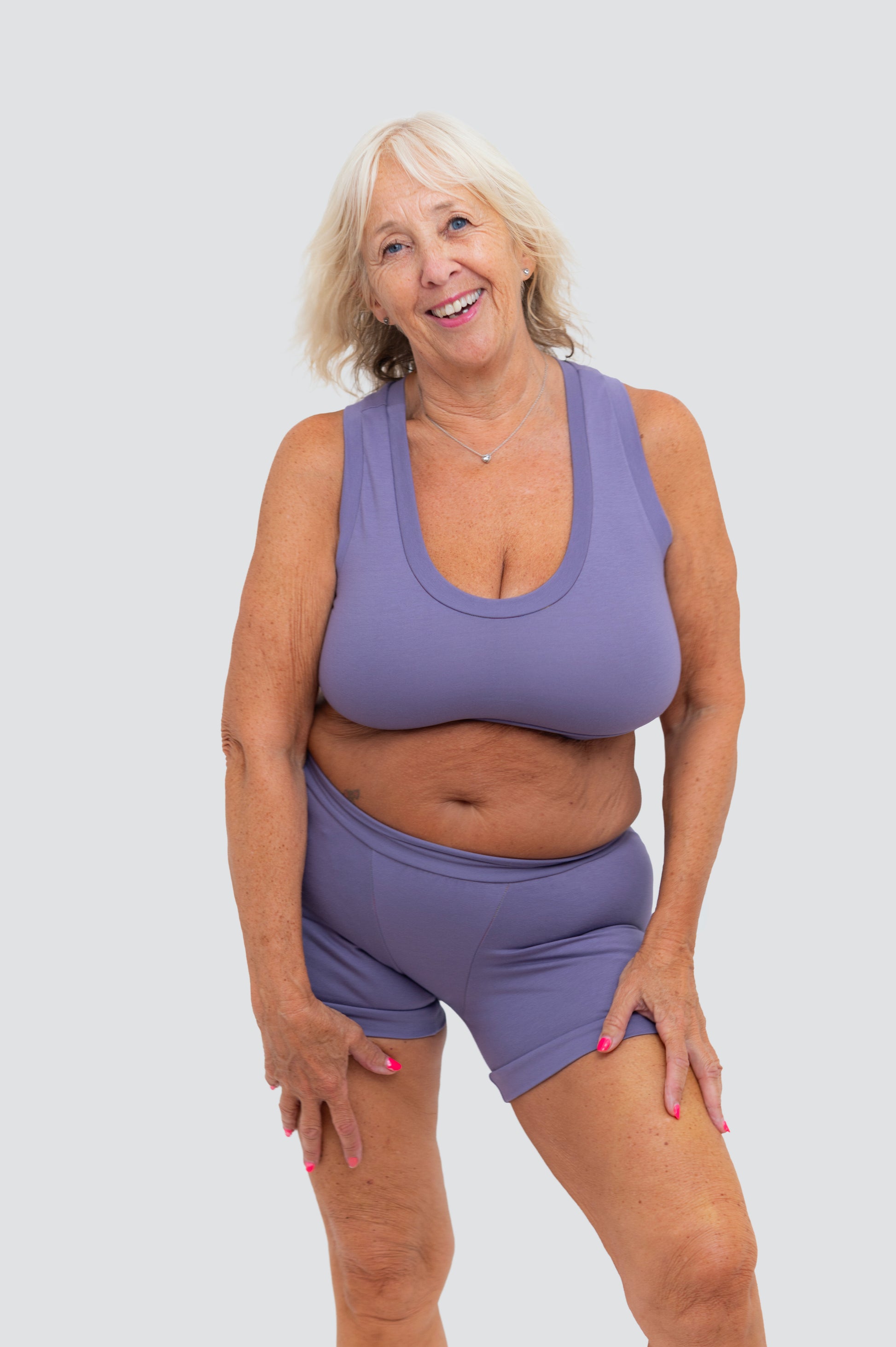 An older woman with blonde hair smiling while wearing a matching lavender-purple athletic tank top and shorts set against a solid light gray background.