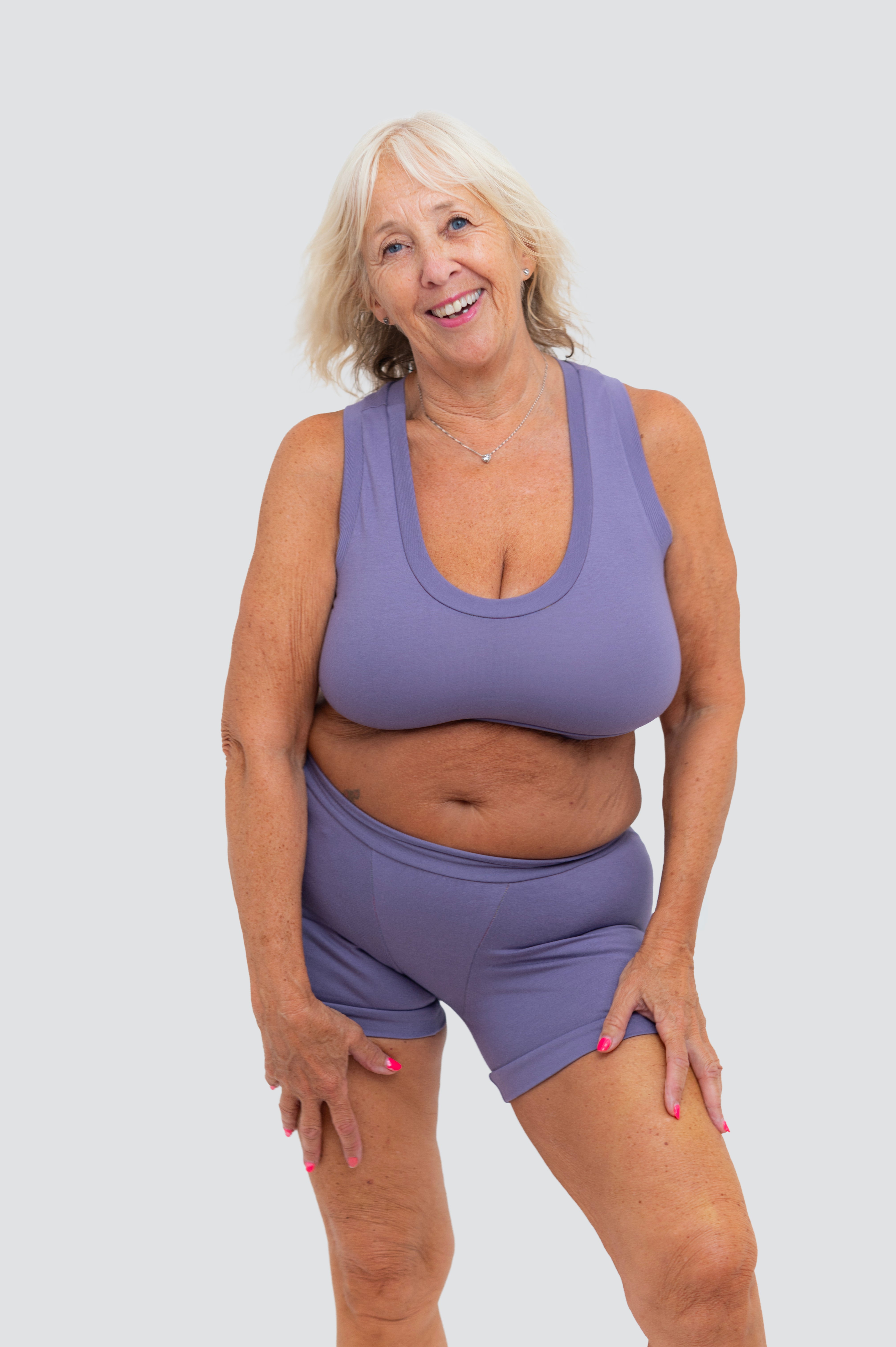 An older woman with blonde hair smiling while wearing a matching lavender-purple athletic tank top and shorts set against a solid light gray background.
