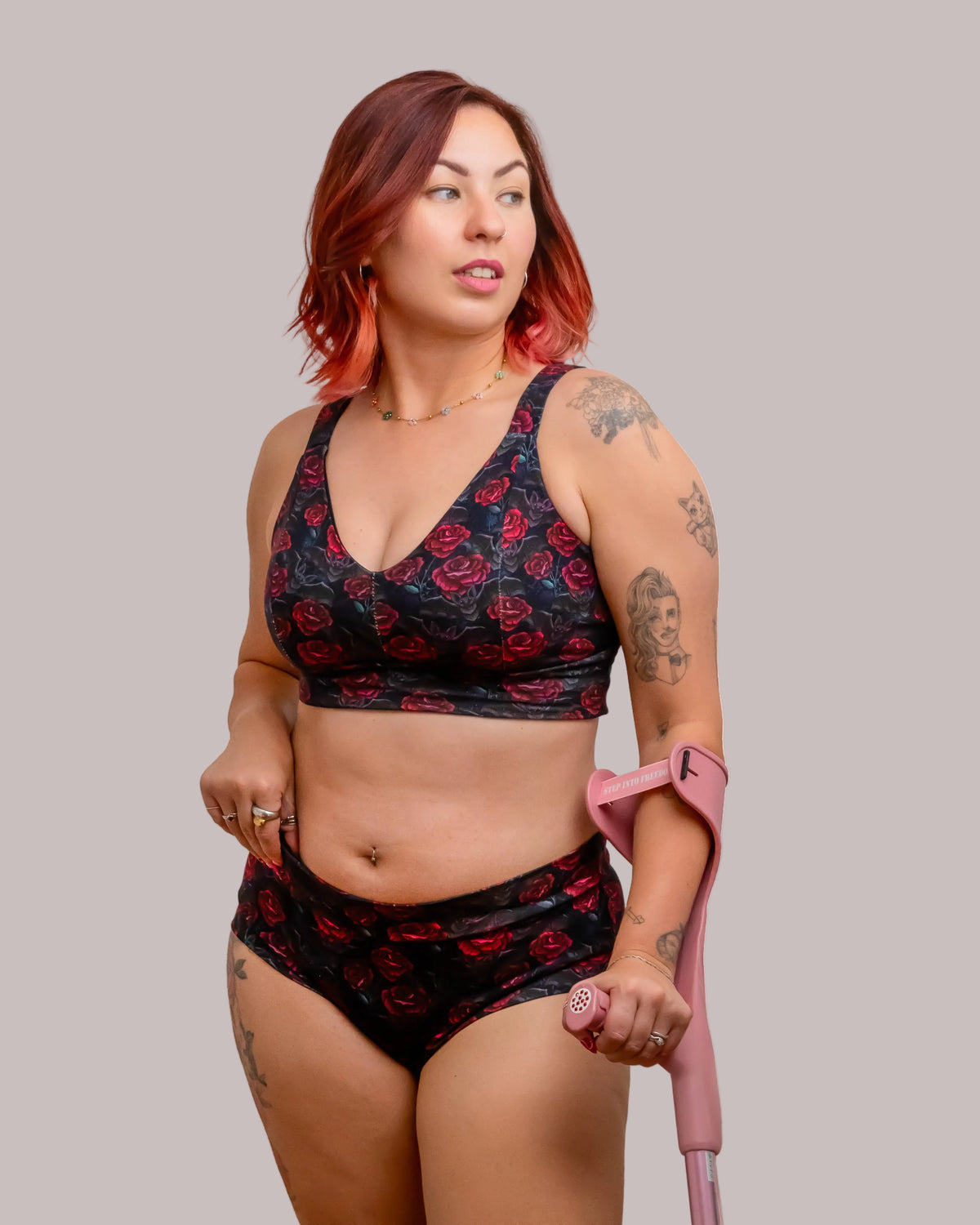 A woman with red hair and tattoos models a matching black and red rose print bra and high-waisted brief set, holding a pink walking stick against a white background.