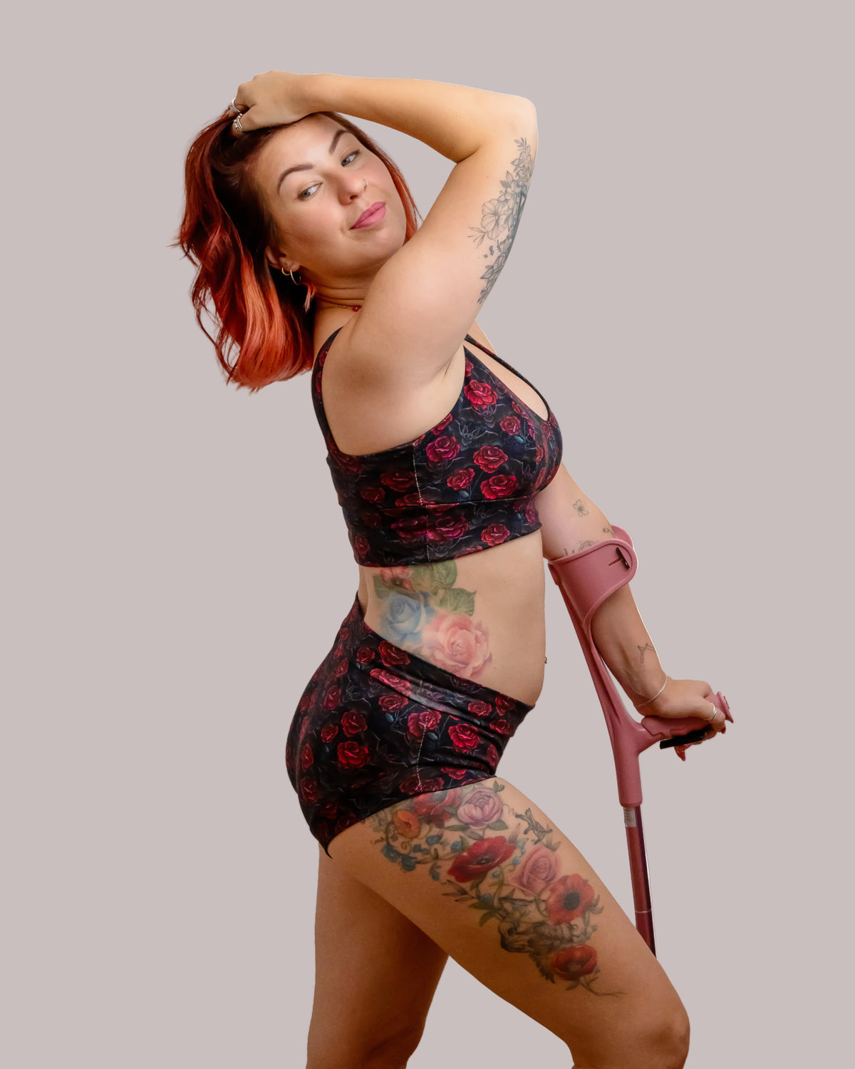 A woman with red hair and tattoos poses in a matching black and red rose print activewear set, featuring a sports bra and high-waisted shorts, while holding a pink crutch against a solid white background.