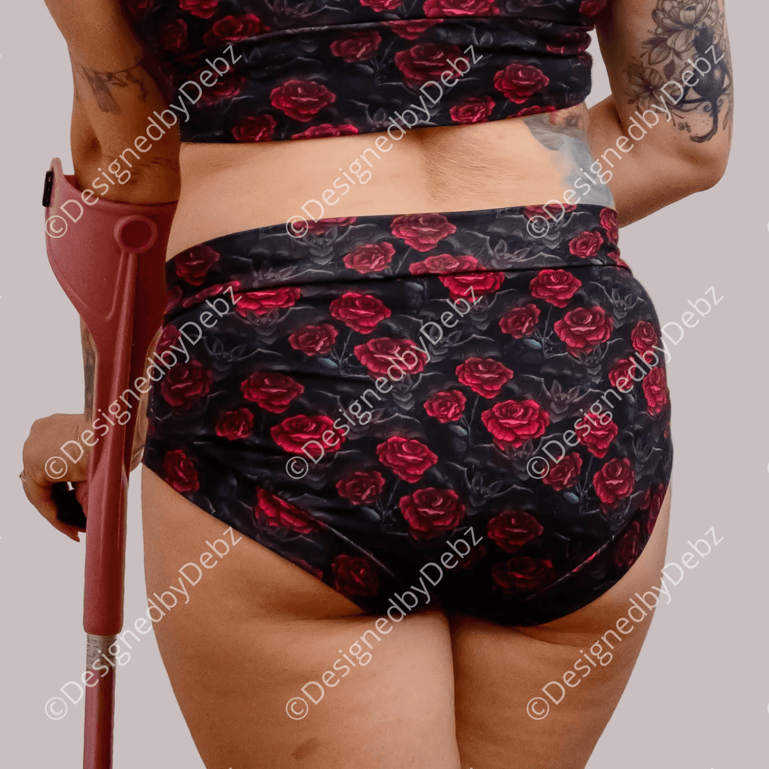 Rear view of a person wearing a matching black floral bikini or swim set with red rose print, holding a reddish crutch against a plain white background.