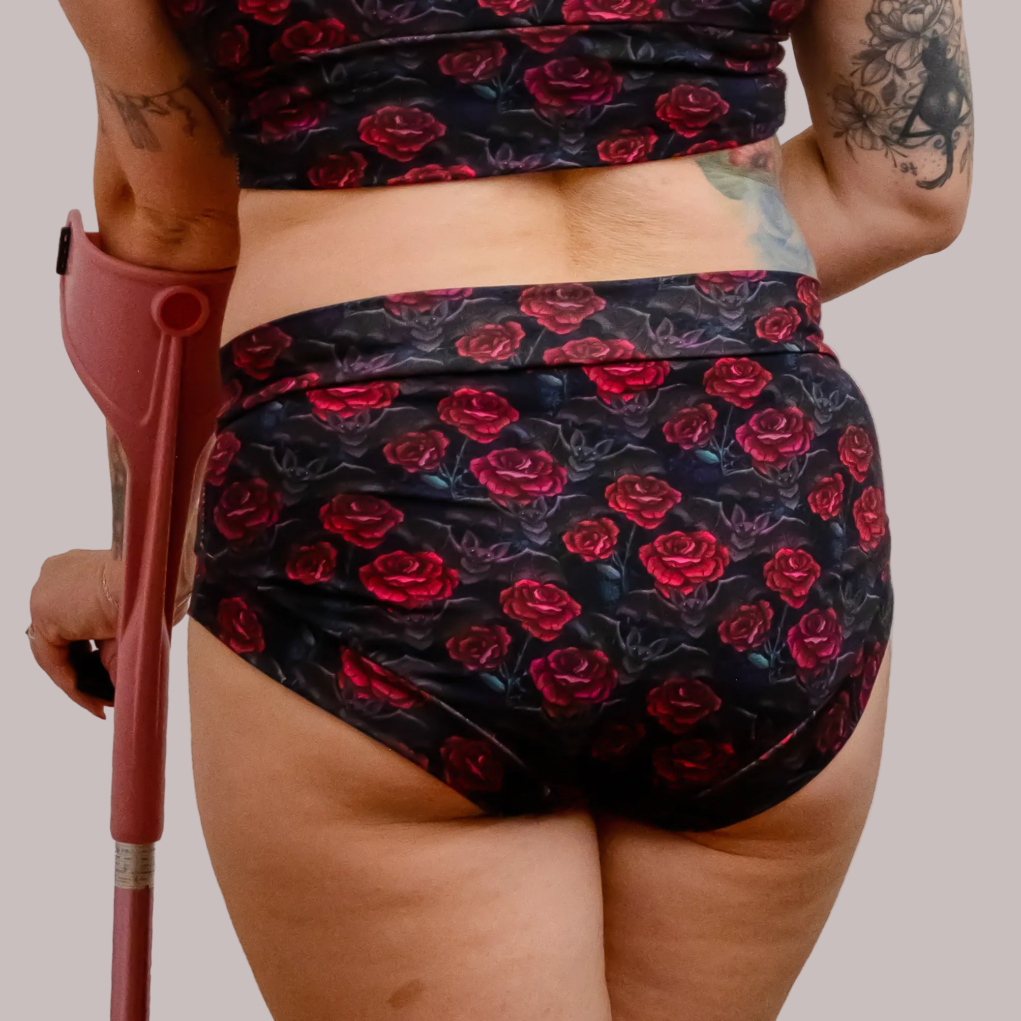 Rear view of a person wearing a matching black floral bikini or swim set with red rose print, holding a reddish crutch against a plain white background.