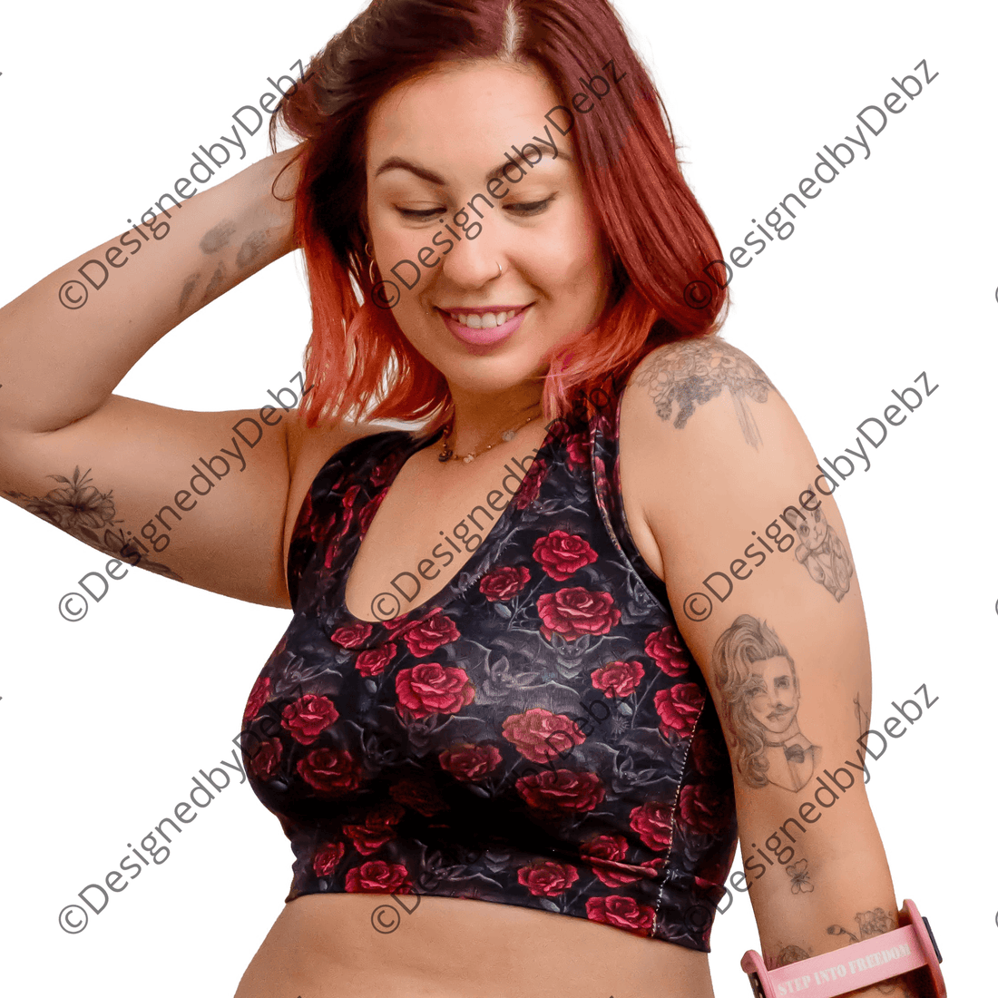 A woman with red hair and tattoos models a matching black and red rose print activewear set, consisting of a sports bra and high-waisted shorts, while holding a pink crutch against a solid white background.