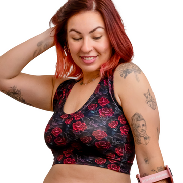 A woman with red hair and tattoos models a matching black and red rose print activewear set, consisting of a sports bra and high-waisted shorts, while holding a pink crutch against a solid white background.