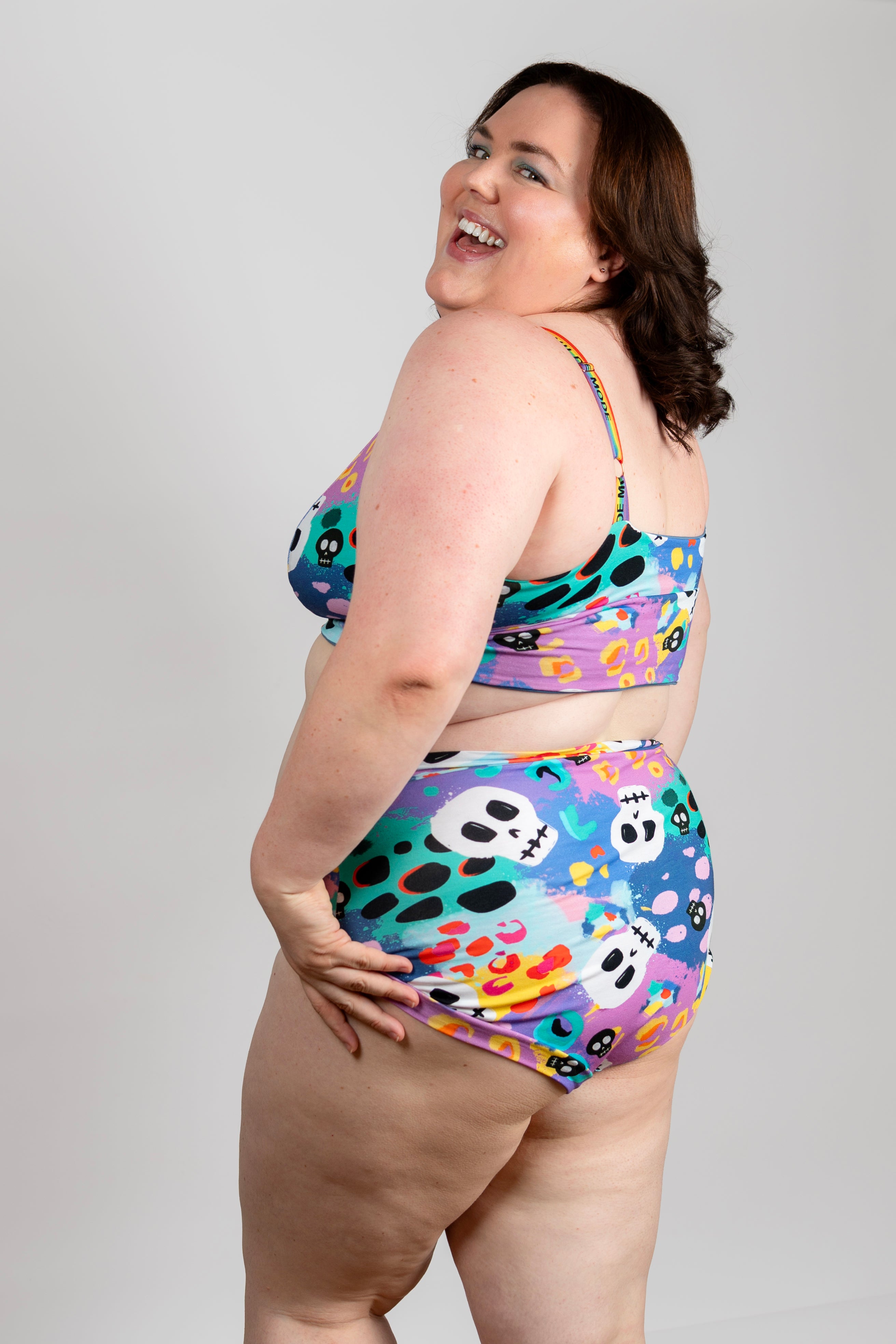 A plus-size woman modeling a two-piece bikini with a colorful abstract print featuring purple, teal, yellow, pink, and black shapes, photographed against a light gray studio background.