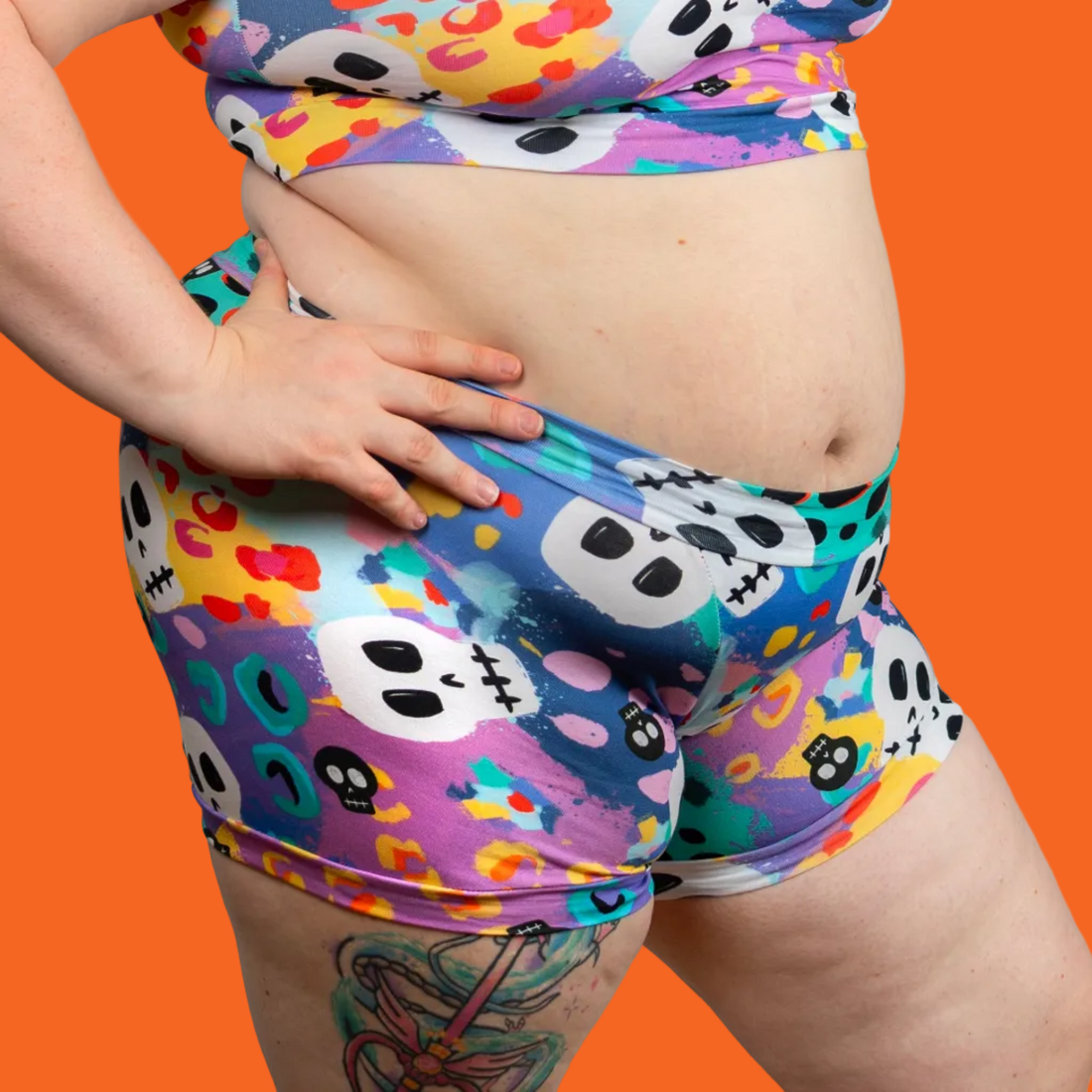 A woman modeling a matching two-piece activewear set featuring a colorful, playful print of skulls, hearts, and dots on a blue base, set against a plain light gray background.