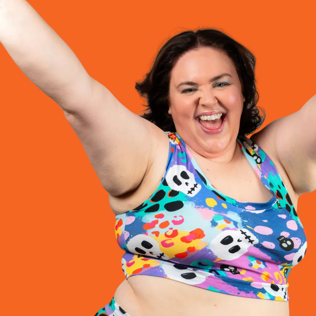 A woman wearing a colorful printed sports bra and matching high-waisted leggings with a playful skull and dot pattern, posing with her arms raised against a solid gray background.
