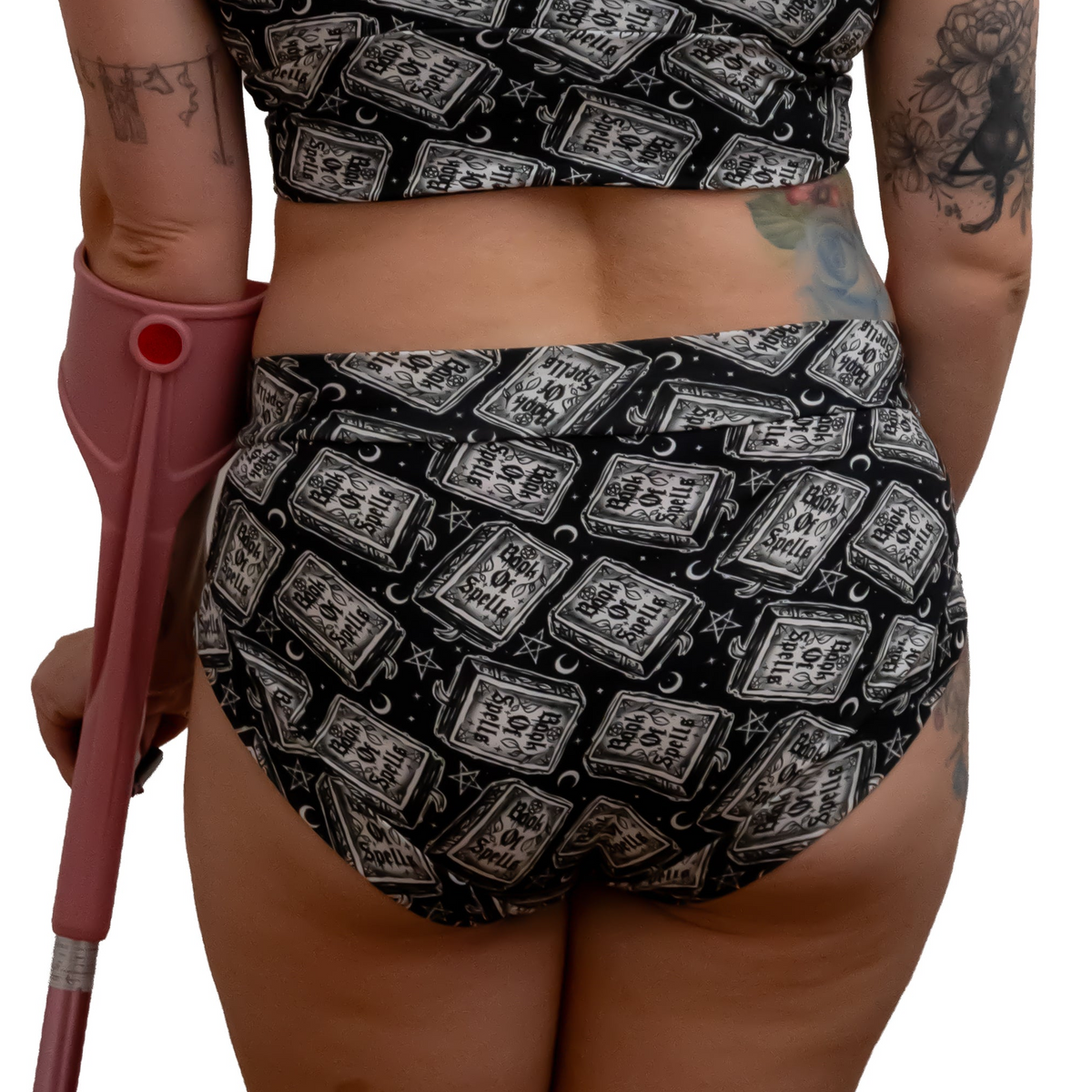Rear view of a woman wearing a black two-piece bikini with a white illustrated pattern of old books and moons, holding a pink crutch against a white background.