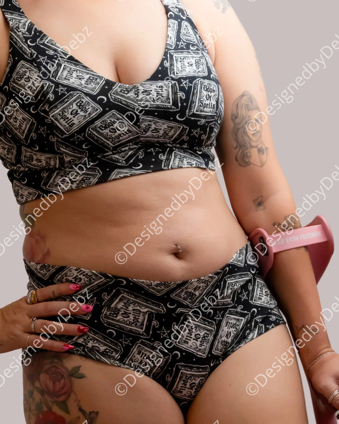 A close-up of a model wearing a matching black and white book of spells  print sports bra and bikini bottom set, with a pink arm strap visible on one arm, holding a pink walking stick