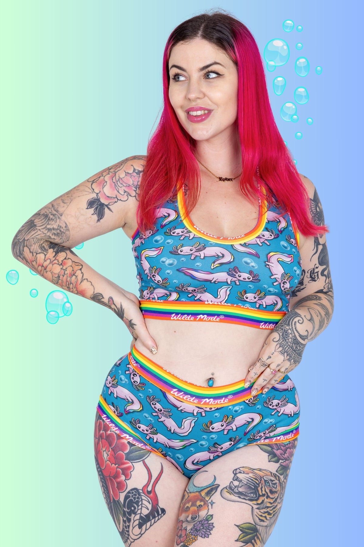 A woman with bright pink hair and tattoos models a matching two-piece swimsuit featuring a blue background with a pink axolotl print and rainbow trim. The background is a soft gradient of green to blue with floating bubble graphics.