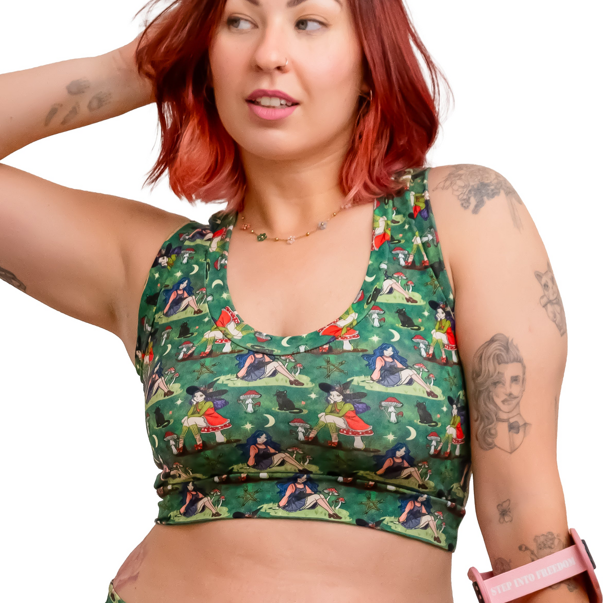 A woman with red hair models a matching green forest witch print activewear set consisting of a sports bra and shorts, holding a pink crutch against a solid white background.