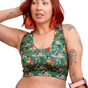 A woman with red hair models a matching green forest witch print activewear set consisting of a sports bra and shorts, holding a pink crutch against a solid white background.