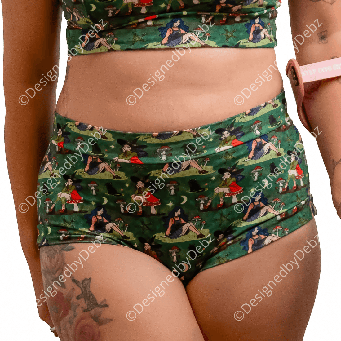 A woman with red hair wearing a green forest witch with cats-print two-piece set consisting of a bra and high-waisted briefs, holding a pink crutch against a solid white background.