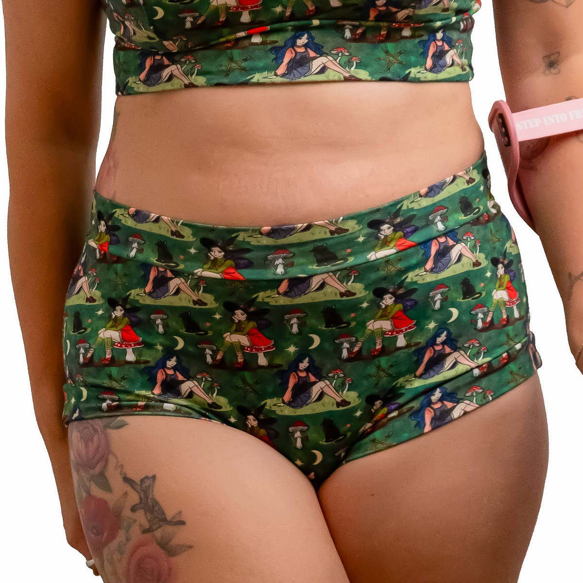 A woman with red hair wearing a green forest witch with cats-print two-piece set consisting of a bra and high-waisted briefs, holding a pink crutch against a solid white background.