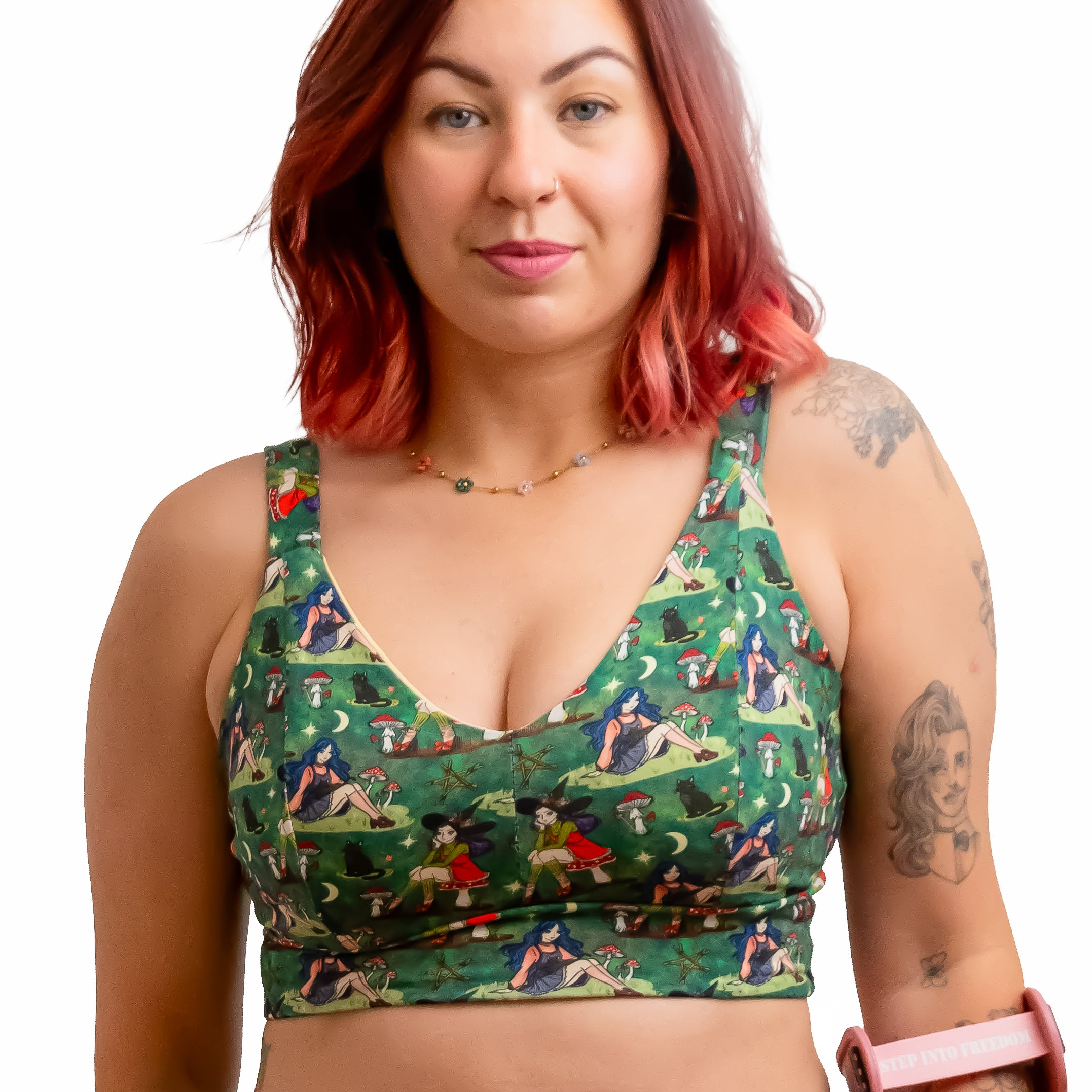 A woman with red hair wearing a green witch-print two-piece set consisting of a sports bra and high-waisted briefs, holding a pink crutch against a solid white background.