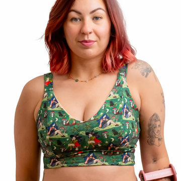 A woman with red hair wearing a green witch-print two-piece set consisting of a sports bra and high-waisted briefs, holding a pink crutch against a solid white background.