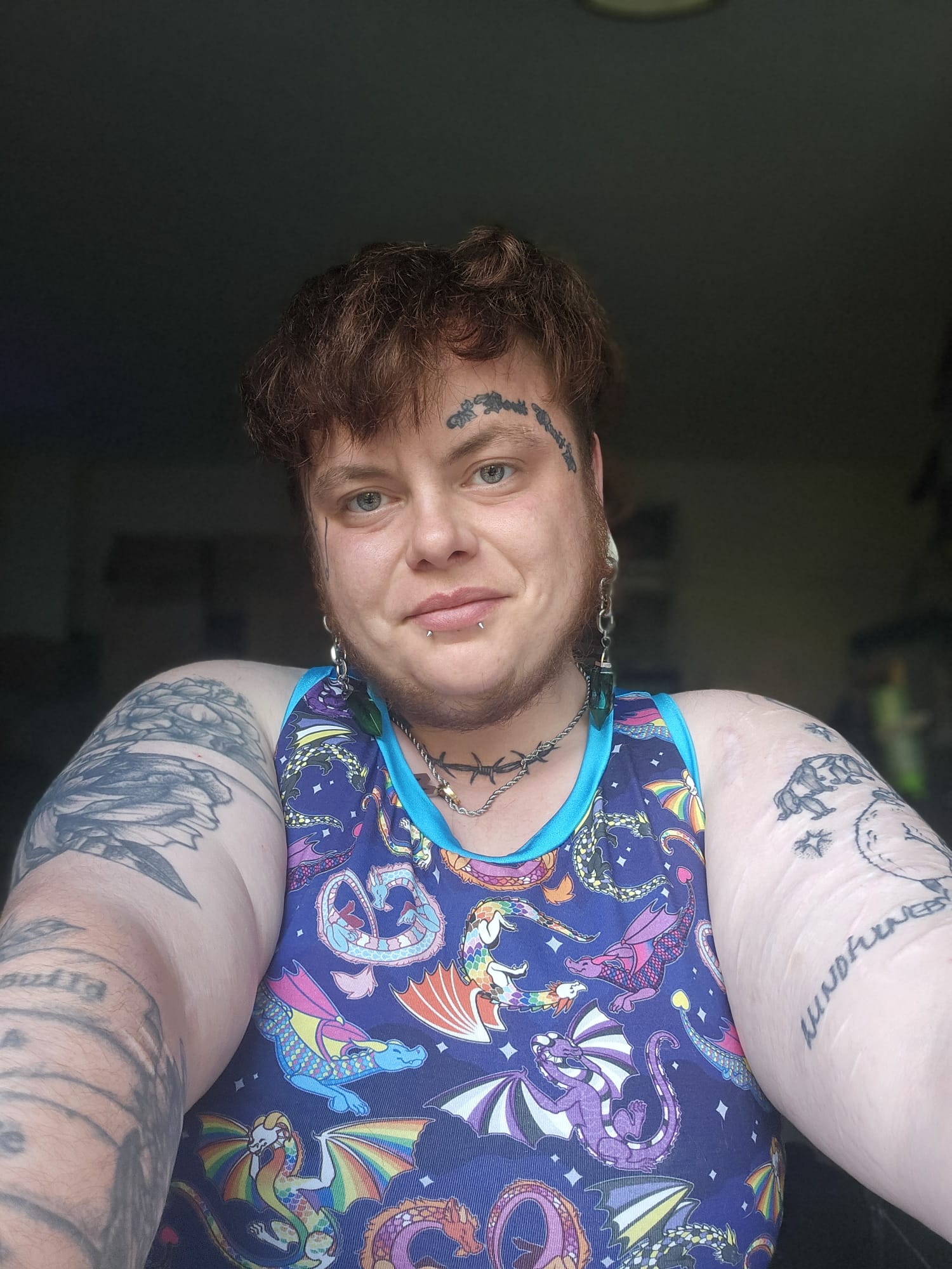 A person taking a selfie wearing a sleeveless binder compression top with a colorful cosmic print featuring dragons, stars, and planets on a dark blue background. The person has short brown hair, visible tattoos on both arms, and is wearing silver hoop earrings and a black choker necklace.