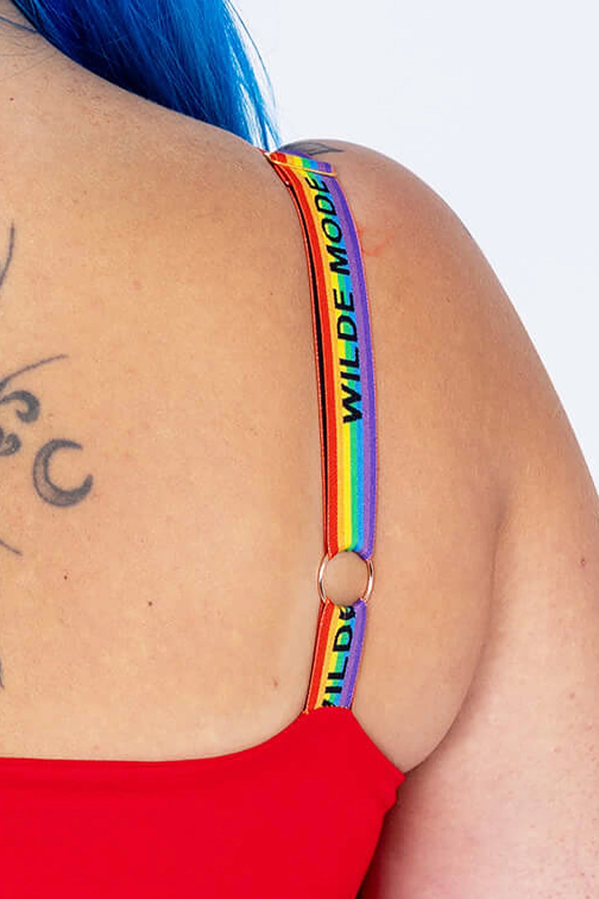 Close-up of a person wearing a rainbow-striped shoulder strap with black text reading 'WILDE MODE' and a gold ring connector, paired with a red top against a light gray background.