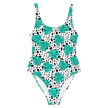 A one-piece swimsuit with a white base, decorated with a pattern of teal monstera leaves and black irregular spots, shown against a plain white background.