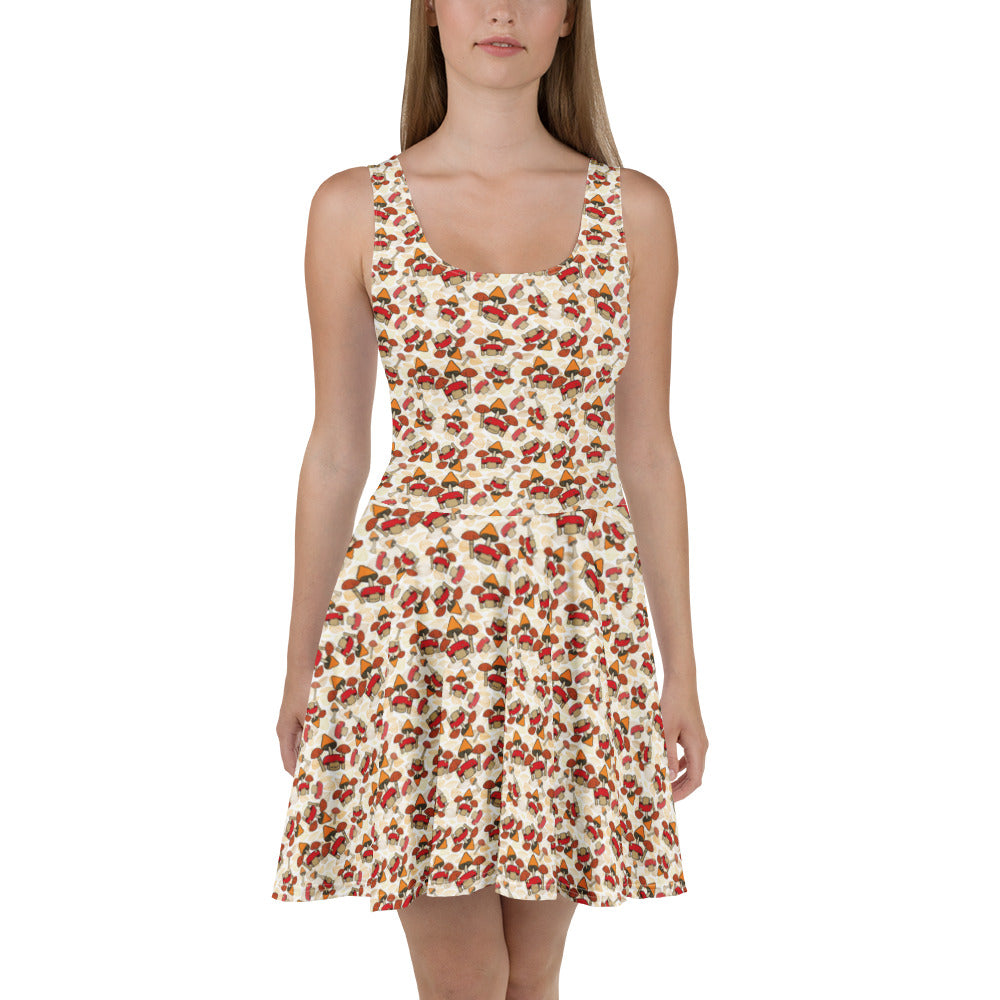 A woman modelling a sleeveless fit-and-flare dress with a warm cream background and an allover small floral print in red, orange, and green mushrooms and toadstools shown against a plain white studio background.