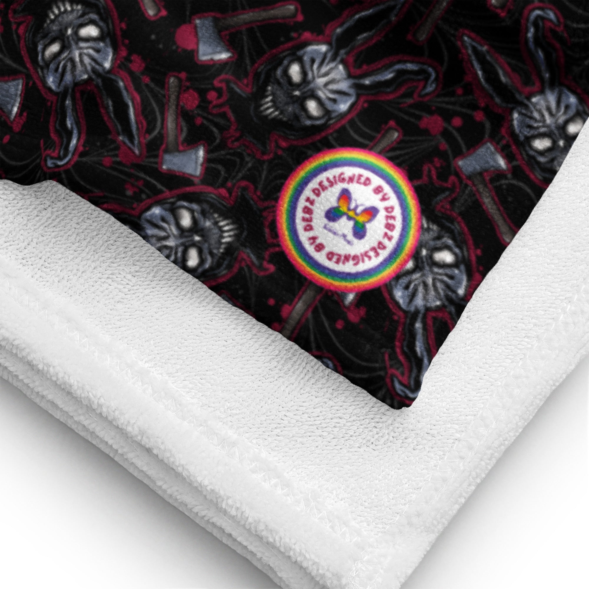 A folded white plush blanket with a black printed backing featuring skulls, bats, and axes, plus a colorful circular patch reading 'DESIGNED BY DESIGNS ALLIGED BY'.