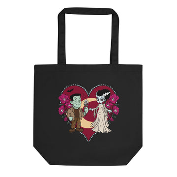 A black canvas tote bag with a cartoon graphic of a vampire and a zombie holding a crescent moon inside a red heart, surrounded by pink flowers, on a plain white background.