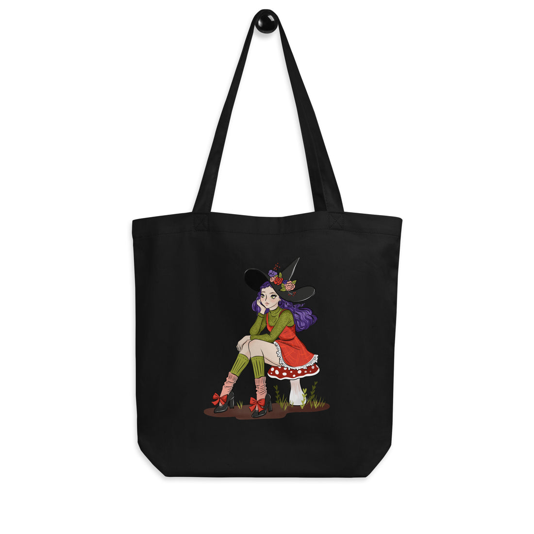 A black canvas tote bag with two shoulder straps, featuring a colorful graphic print of a witch sitting on a rock. The bag is displayed against a clean white background.